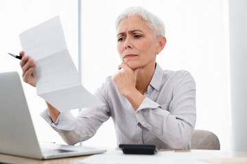 Confused Caucasian mature businesswoman paying bills using laptop at office. Middle-aged manager CEO HR analyzing reading bank statement, mortgage document, formal letter