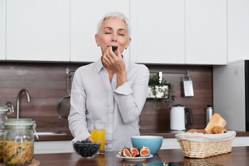 Happy mature woman eating figs for breakfast with oat flakes and granola in the morning. Middle-aged housewife eating healthy food at home kitchen