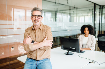 Senior leader stands with arms crossed in front of glass partition, a colleague working in the background, suggesting a supportive yet focused work environment that values leadership and concentration