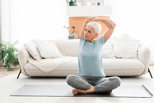 Caucasian female athlete stretching her arms in yoga lotus position at home. Mature middle-aged woman practicing training indoors on fitness mat