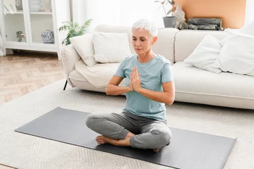 Tuinposter Cheerful Caucasian mature woman in fitness clothes meditating in lotus position while doing yoga at home. Middle-aged female athlete practicing home workout © InsideCreativeHouse