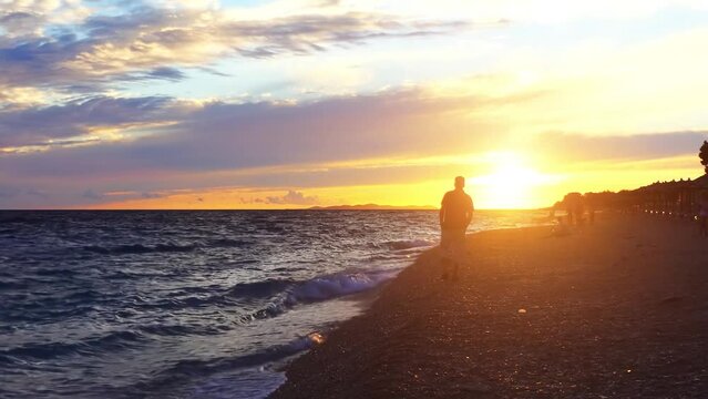 Lone man goes along pebble beach. Evening sun sits in sea. Resort town on shores of Adriatic Sea.