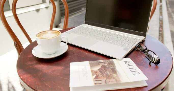 A laptop, a cup of coffee, glasses, and a magazine rest on a cafe table