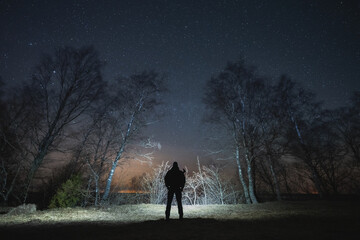 Silhouette of a man with a headlamp with a rifle behind his back in a night forest under a starry sky. © Dmitri