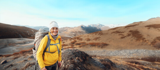 A smiling senior woman in a yellow jacket and white beanie stands on a mountain trail, her face alight with the joy of adventure. Banner. Concept travel alone, outdoor activities, vacation, tourism.