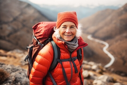 A smiling senior woman in a red puffer jacket and beanie stands on a mountain trail, her face alight with the joy of adventure. Concept travel alone, outdoor activities, vacation, tourism, lifestyle.