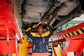 Portrait of an African female mechanic in yellow and blue uniform standing under the car bottom for inspecting in garage. Woman smiling while holding a wrench. Car repair service. Vehicle maintenance - 757280673