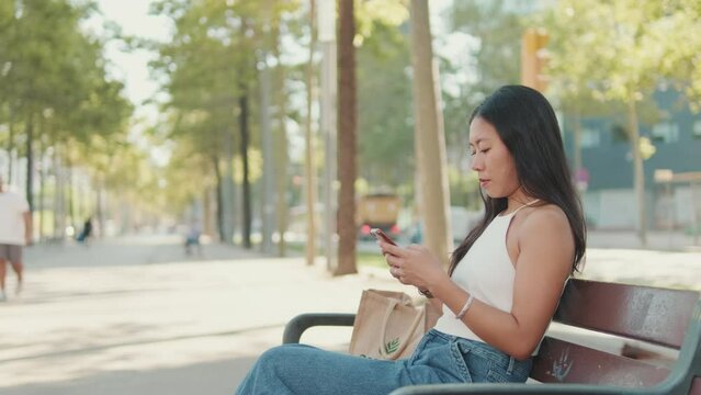 Young woman sitting on park bench typing message on mobile phone