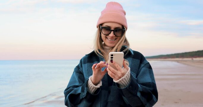smiling woman using mobile phone while walking on the beach coast