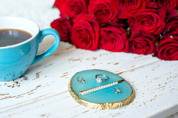 a bouquet of red roses, a cup of coffee and  jewelry on a white tray on a white bed in the bedroom.
