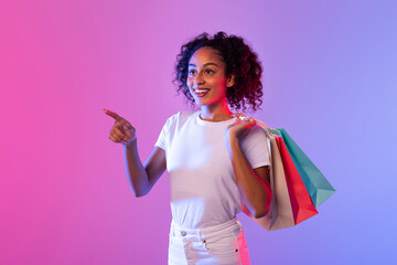 Happy lady shopper pointing away with colorful bags on neon-lit background