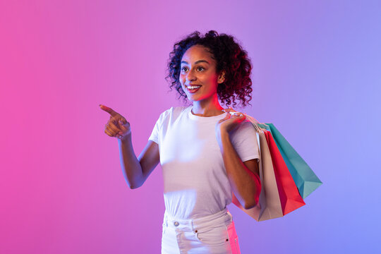 Happy lady shopper pointing away with colorful bags on neon-lit background
