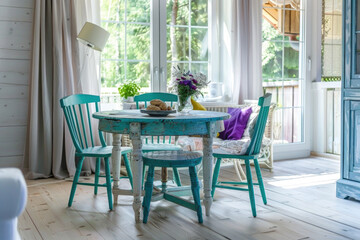 Rustic, vintage furniture, table and chairs of various bright colors in the dining room with large windows.