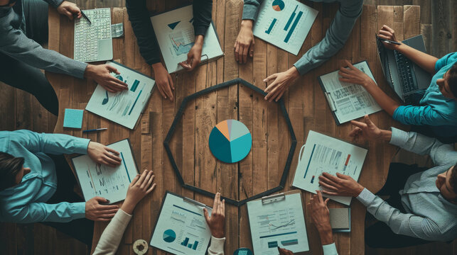 top view of several people's hands pointing and reaching towards a colorful pie chart on a table surrounded by business reports and documents, indicating a collaborative work environment