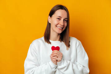 Cute young smiling woman wearing white is holding a red heart at her chest. Studio shot over yellow background.