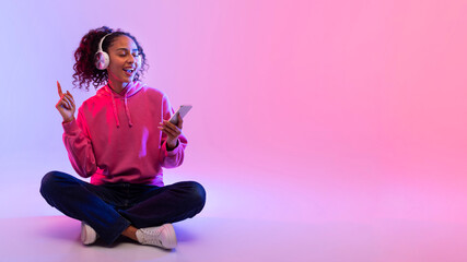 Happy black woman in hoodie enjoying music on headphones, using smartphone