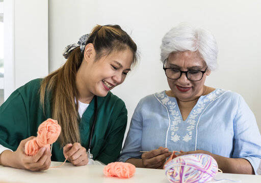 Young Asian care taker teaching eldery Indian woman crocheting in senior homecare