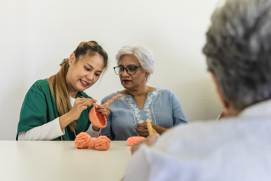 Young Asian care taker teaching eldery Indian woman crocheting in senior homecare