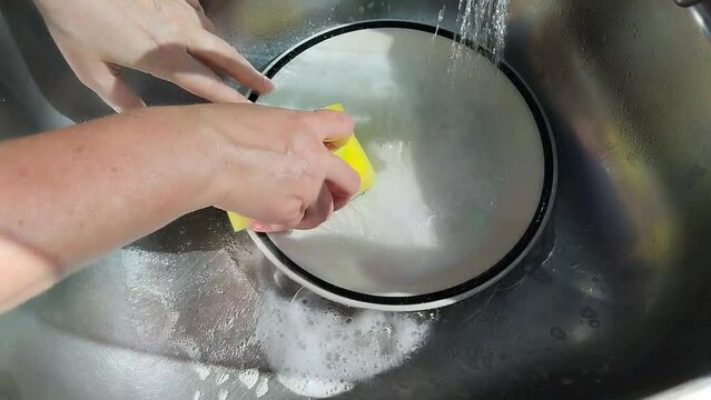 Unas manos de  mujer lavando un plato de color blanco  con una esponja amarilla con jab&oacute;n liquido y agua  en un fregadero