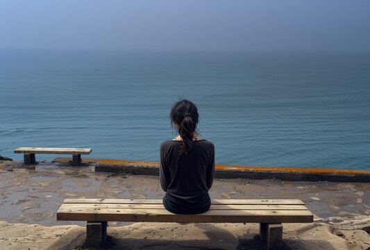 A depressed dishevelled girl sitting on an empty bench overlooking the ocean, back view. 