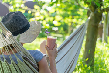 Young woman with hat resting in comfortable hammock at green garden with Glass of fresh strawberry milkshake, smoothie. Healthy food and drink concept.