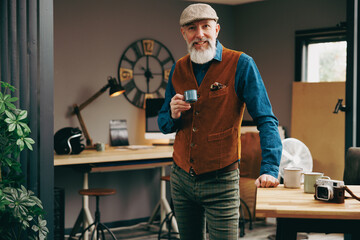 Portrait d'un homme debout souriant quinquagénaire senior hipster élégant et stylé qui boit un café dans un atelier créatif vintage