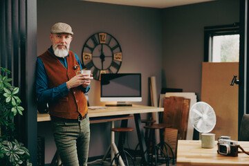 Portrait d'un homme debout souriant quinquagénaire senior hipster élégant et stylé qui fait une pause et qui boit un grand café dans un atelier créatif vintage - 757373681