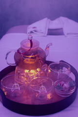 Glass teapot with herbal tea and candle on massage couch. Spa salon interior. Luxury lifestyle. Thermal centre. Teapot with cups on tray in purple light. Recreation and aroma treatment.  - 757376066
