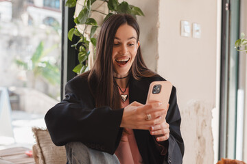 Portrait of happy female dressed casually holding mobile phone, typing messages, communicating with friends via social networks, using high Internet connection at cafe. Woman make video call, wow.