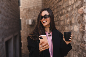 Beautiful smiling woman walking on street with coffee takeaway, chatting on mobile phone. Girl talking on smartphone on her way to meeting spot, sending message, wear black suit and black glasses.