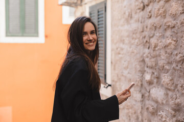 Photo of charming pretty young lady hold modern gadget smiling outside urban city street. Pensive brunette woman student freelancer wear black jacket walking in old town, hold phone in hand.