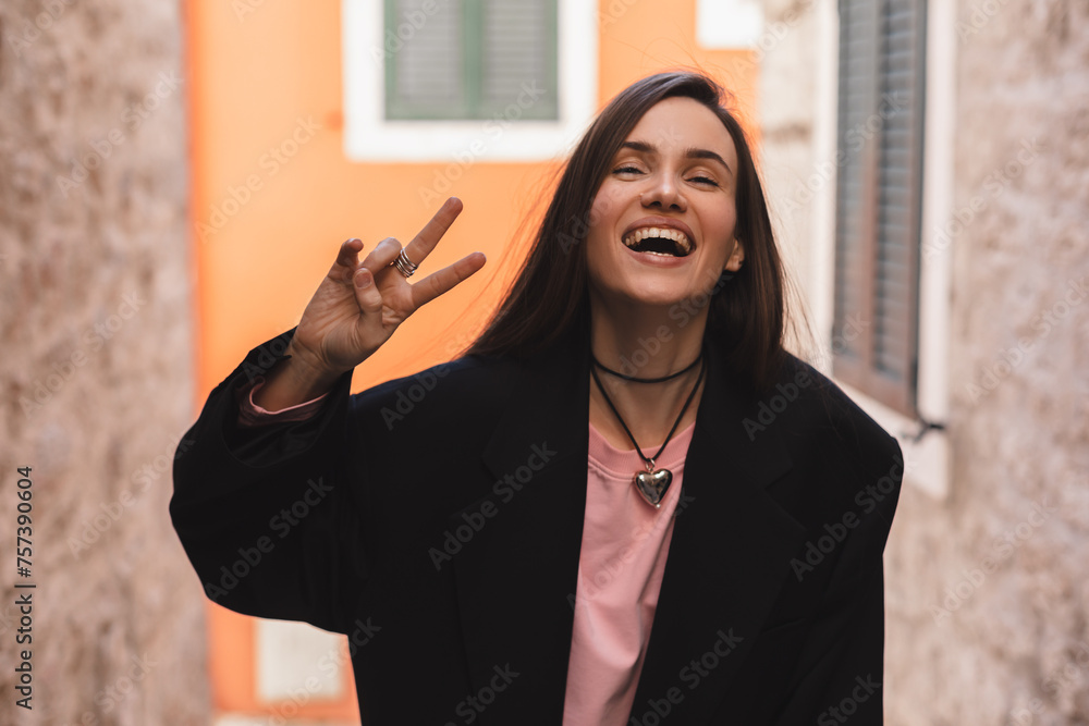 Canvas Prints Cool brunette woman in black jacket, pink long sleeves top and heart shape necklace shows sign v, peace and friendship, open mouth and posing outside. Happy woman walk and posing outdoor in the city. 