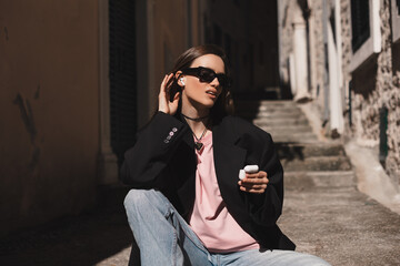 Smiling young woman sitting on stairs and putting wireless earphones on. Woman is relaxing listening music in old town. Young woman using technologies, wear black glasses, black jacket, hold case.