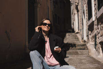 Smiling young woman sitting on stairs and putting wireless earphones on. Woman is relaxing...