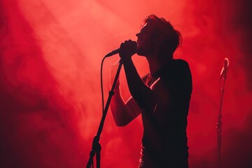 A man singing into a microphone on stage in front of a red background. The man is wearing a black shirt and black pants - obrazy, fototapety, plakaty