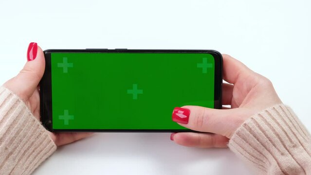 Female hands with green screen chroma key smartphone in horizontal position close up on white background. Woman with red nails holding phone, touching, swiping