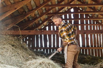 cheerful good looking man with tattoos using pitchfork while working with hay while on farm