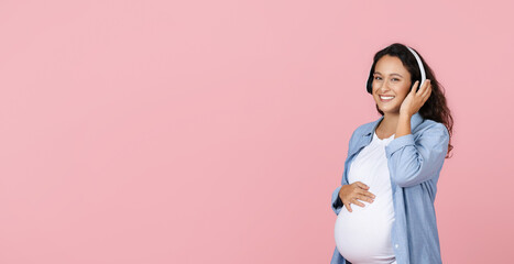 Pregnant woman listening music in headphones on pink background