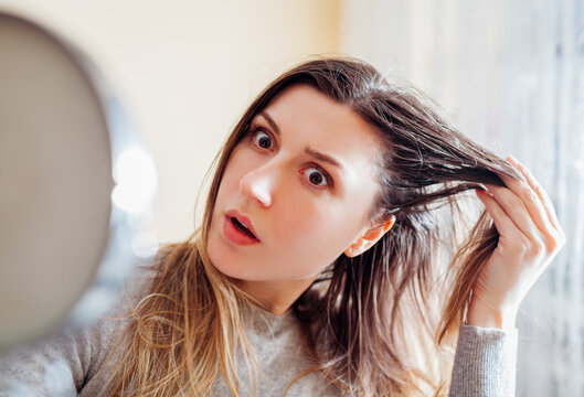 Young shocked woman is surprised to see dirty oily and greasy hair looking in mirror at home. Health treatment