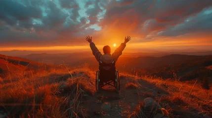 Fotobehang A boy in a wheelchair on a grassy knoll, with his hands raised high, facing a breathtaking sunset that paints the sky and the mountainous horizon in shades of orange © Алексей Василюк