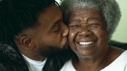 Adult grandson kissing elderly African American grandmother on the cheek in tender affectionate loving moment between two intergenerational family members - obrazy, fototapety, plakaty