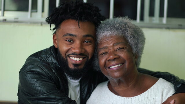 African American young grandson embracing his elderly gray hair grandmother portrait faces looking at camera in genuine loving moment, tender arm around shoulder, intergenerational bond