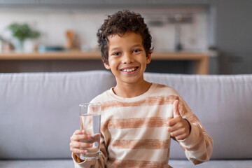 Smiling little black boy holding glass of water and showing thumb up