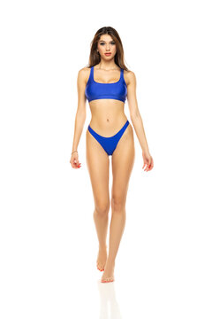 Young beautiful brunette woman in blue bikini swimsuit walking on a white background.