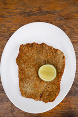 Argentine dish. Top view of milanesa, breaded fried steak, with a slice of lemon, in a white dish with a wooden background.