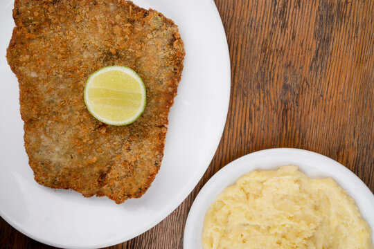 Argentine dish. Top view of milanesa, breaded fried steak, with a slice of lemon and mashed potatoes, in a white dish with a wooden background.	
