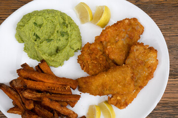 Top view of fried hake fillet with lemon, fried sweet potatoes and beans puree in a white plate with a wooden background.