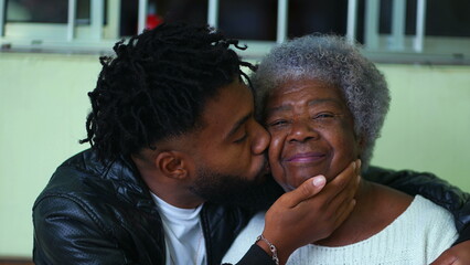 Young African American grandson in 20s kissing elderly Grandmother on the cheek in affectionate caring embrace with arms around senior member of the family, tender intergenerational bond - obrazy, fototapety, plakaty