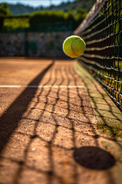 A tennis ball clings to the net casting long shadows on the clay court in the warm sunset light. Generative AI