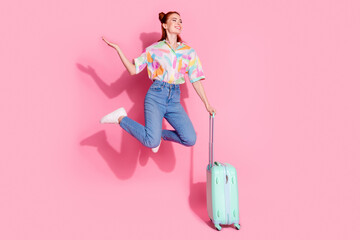 Obraz premium Full size photo of attractive girl colorful blouse jump hold suitcase palm hold offer look empty space isolated on pink color background